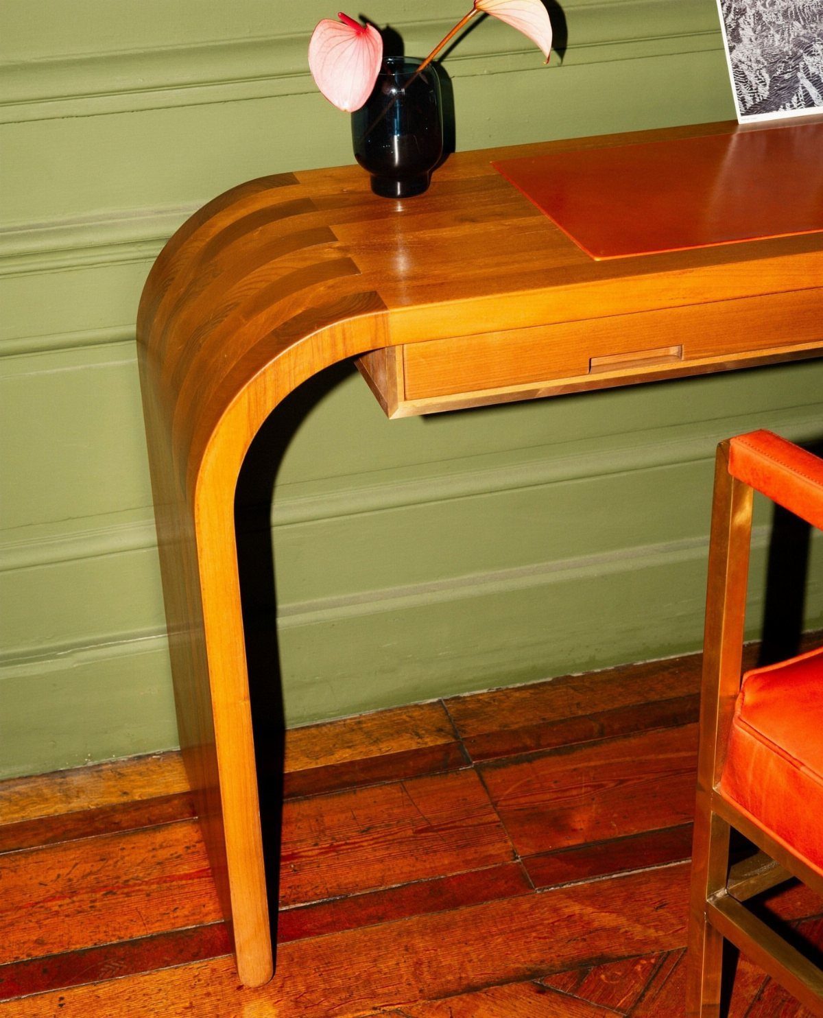 Curved wooden desk with a reddish-orange leather top inlay against a green wall.