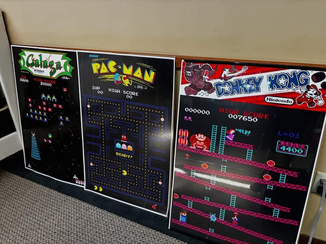 Three retro arcade game posters leaning against a wall on a grey carpet.