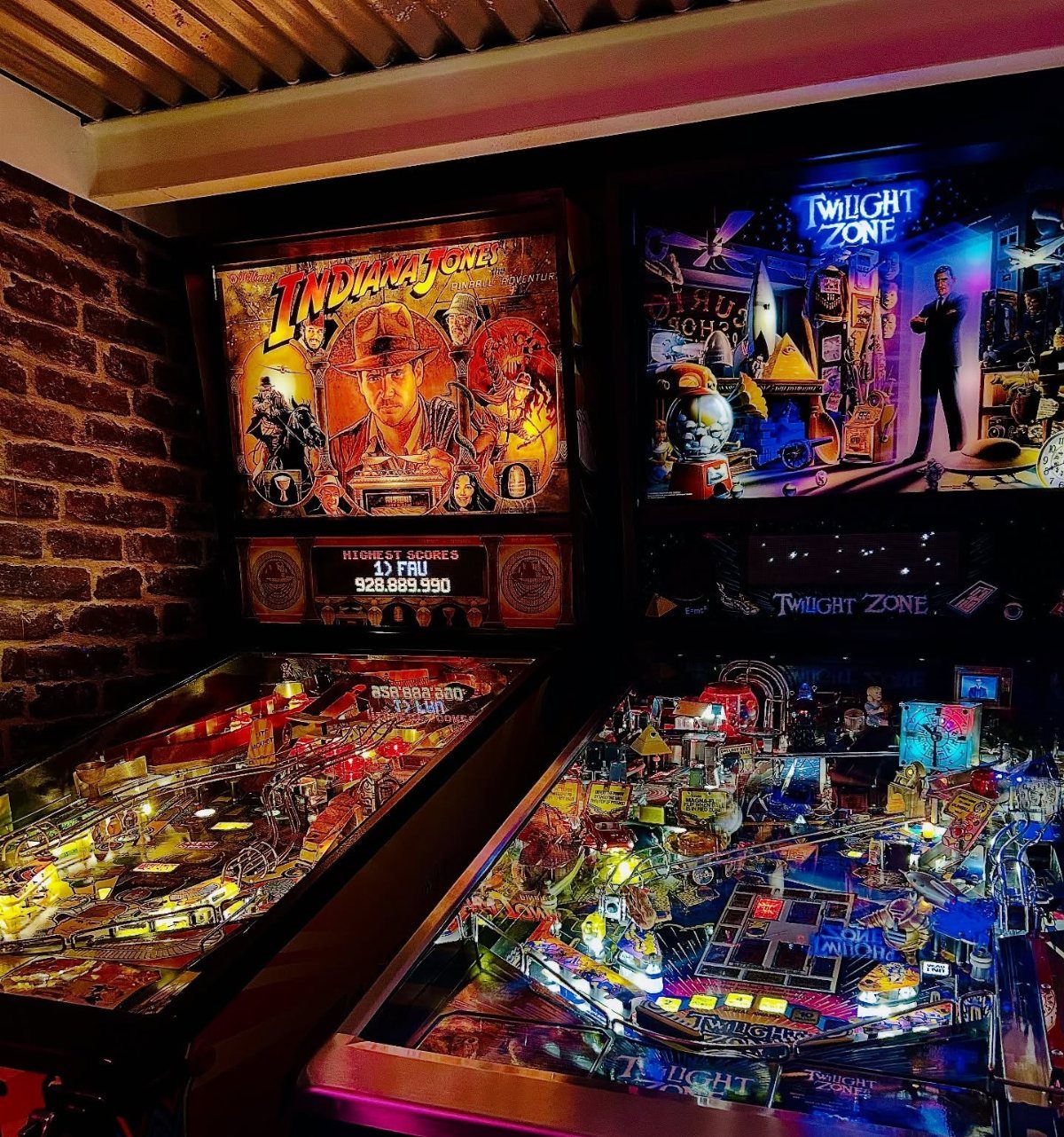 Two vintage pinball machines with glowing displays against a rustic brick wall.