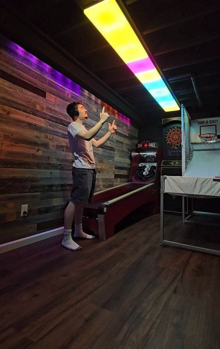 Man in a game room with shuffleboard, basketball hoop, and neon ceiling lights.
