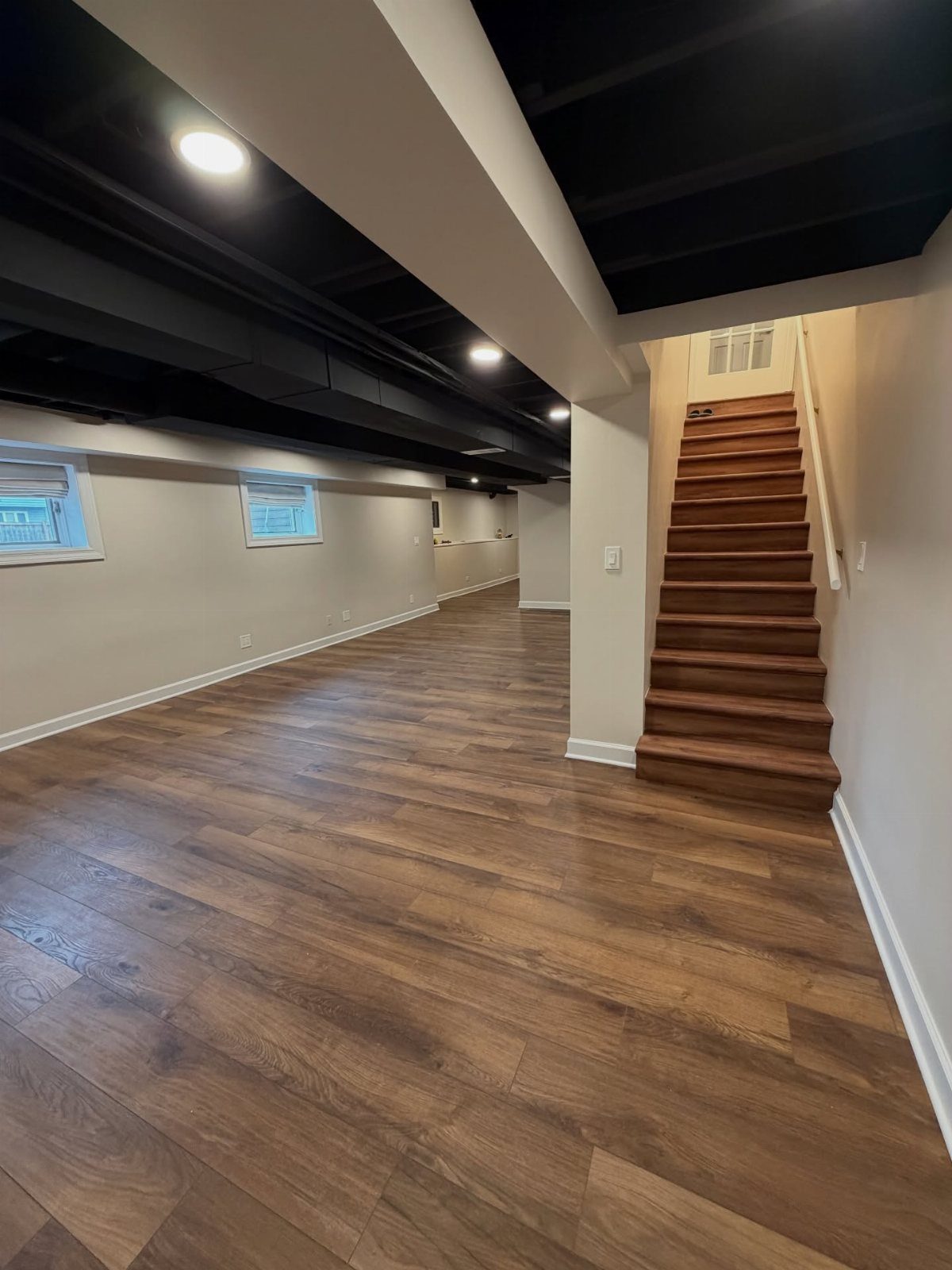 Black painted open-beam basement ceiling with recessed lighting and wood-look flooring.