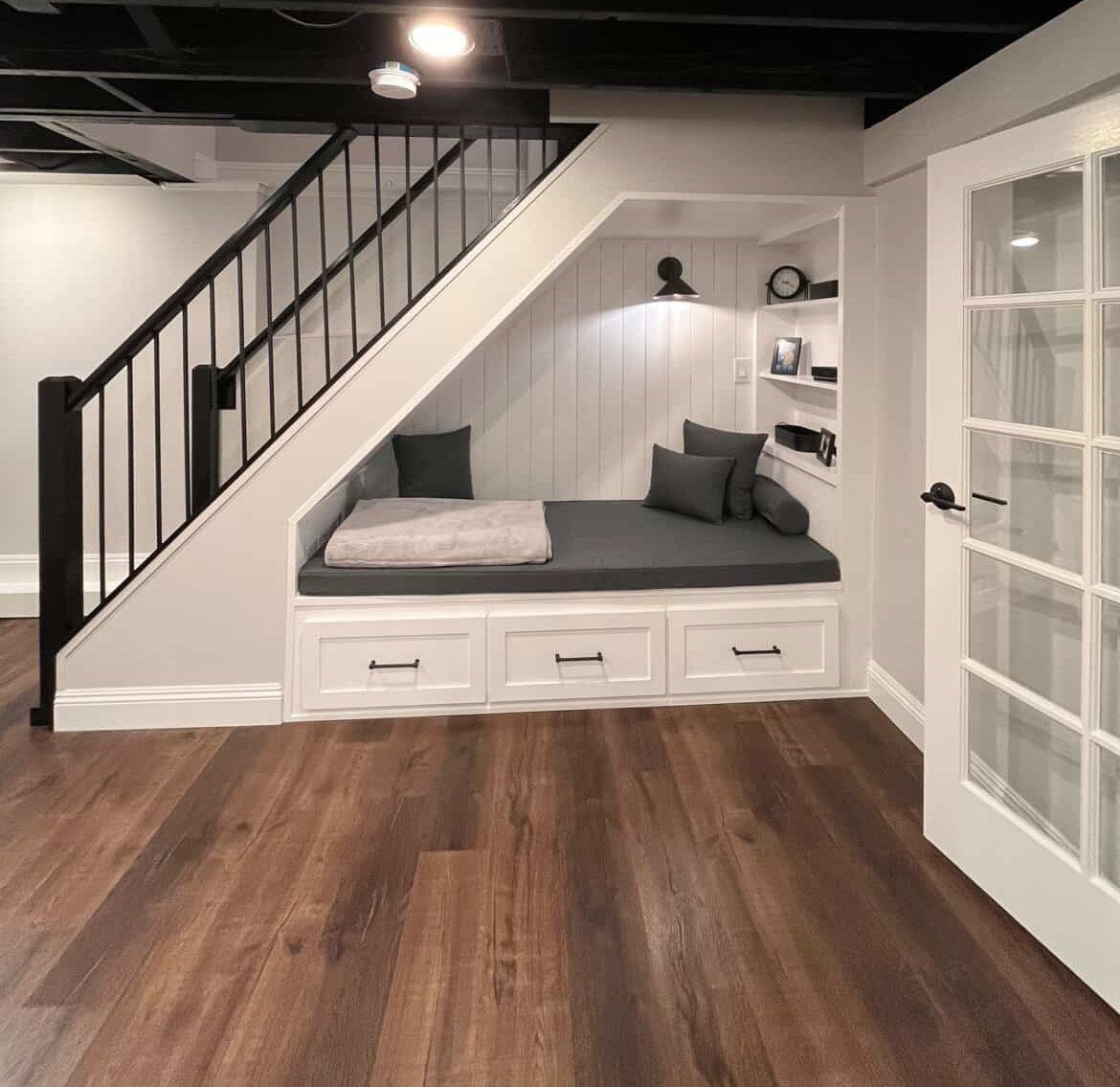 Built-in reading nook with grey cushions under a staircase with black railings.