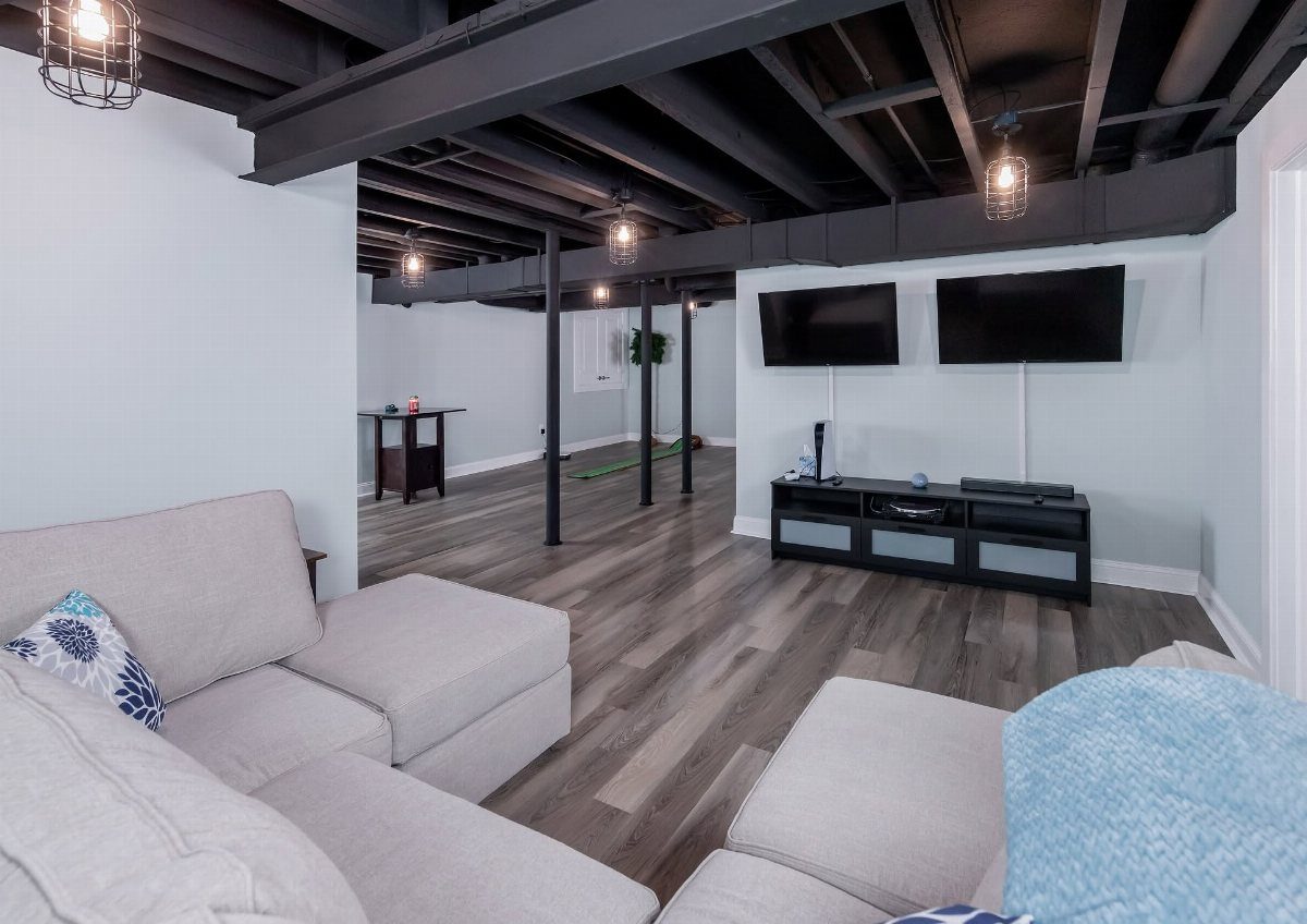 Light grey sectional sofa in a basement with black exposed ceiling and TVs.