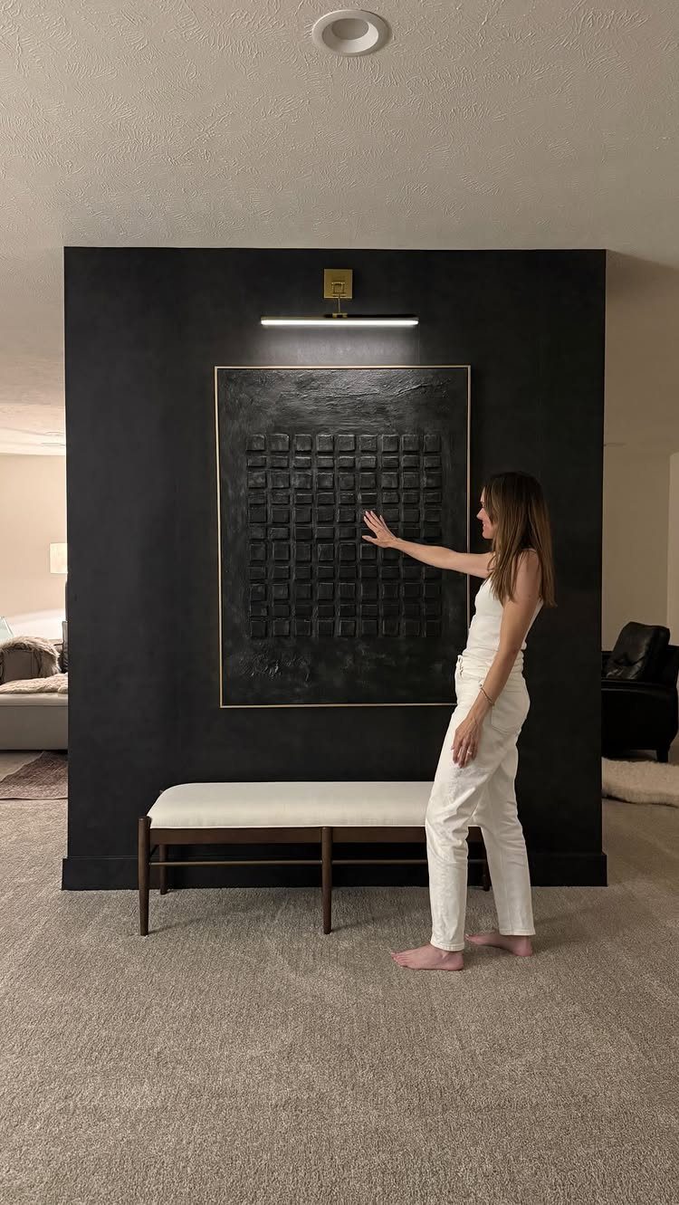 Large textured black art piece on a black wall with a white bench.