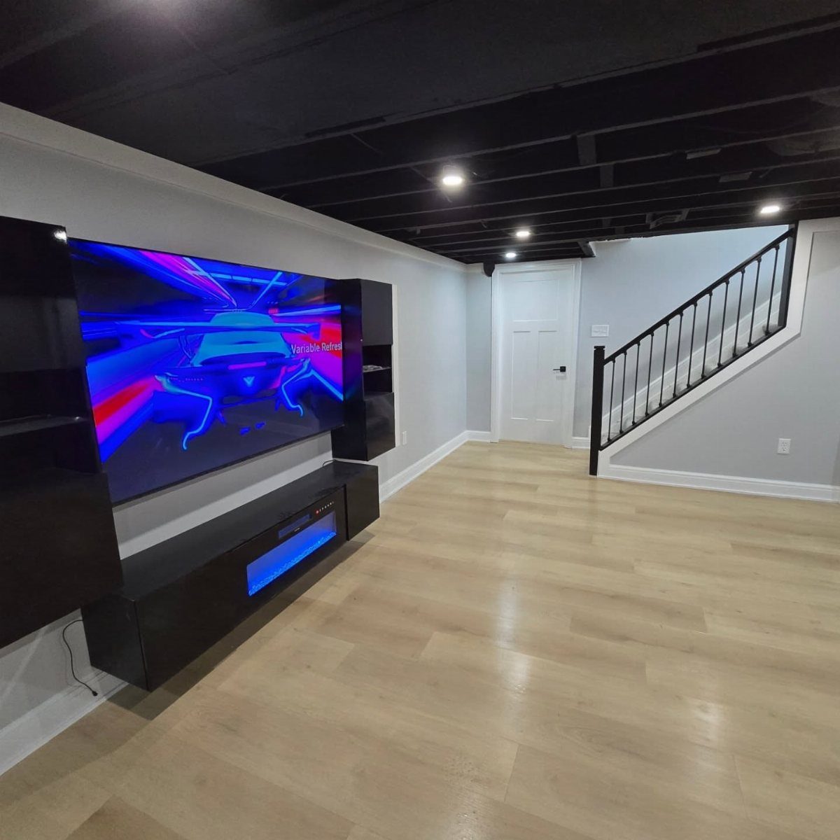 Modern basement living area with a large wall-mounted TV and light wood floors.