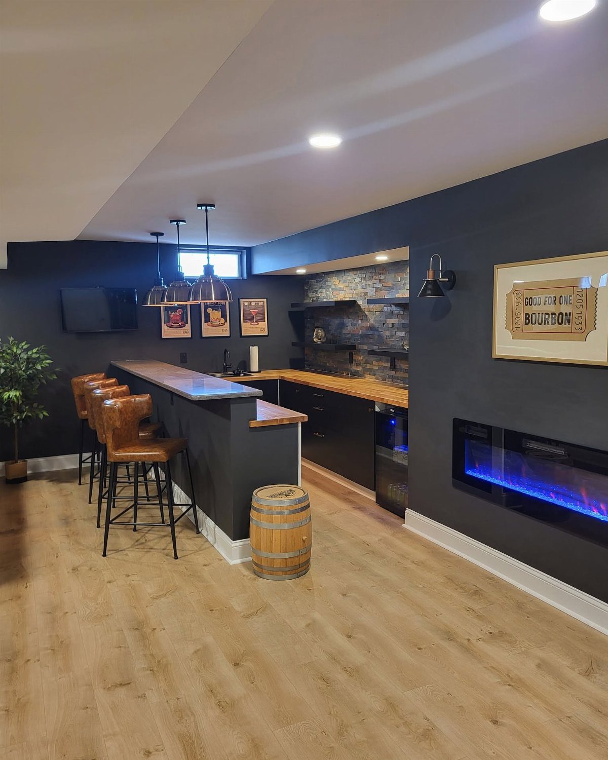 L-shaped basement bar with dark grey cabinets, wooden top, and leather bar stools.