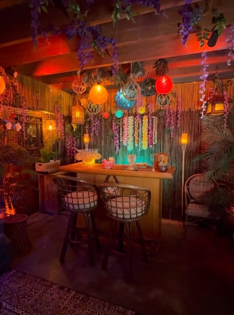 Tiki bar with wooden counter, woven stools, colorful lanterns, and bamboo walls.
