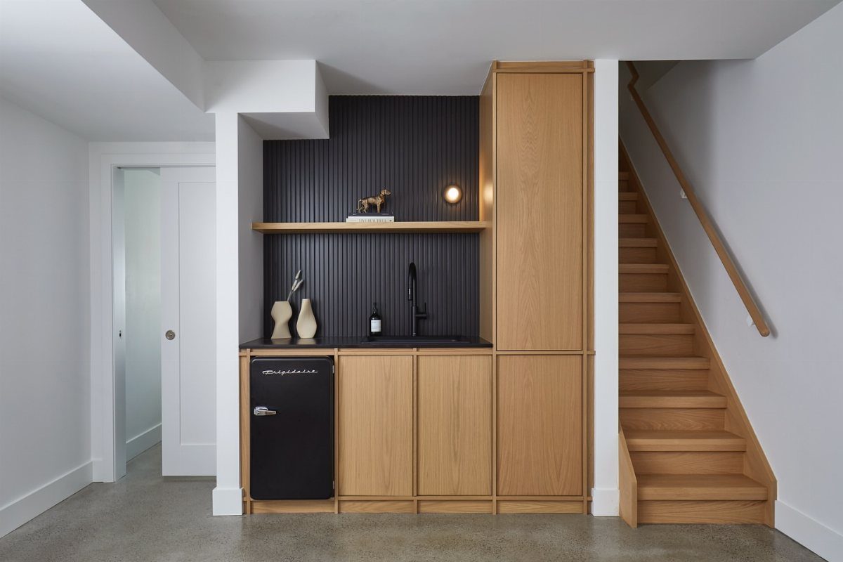 Modern kitchenette under stairs with light wood cabinets and a black mini fridge.