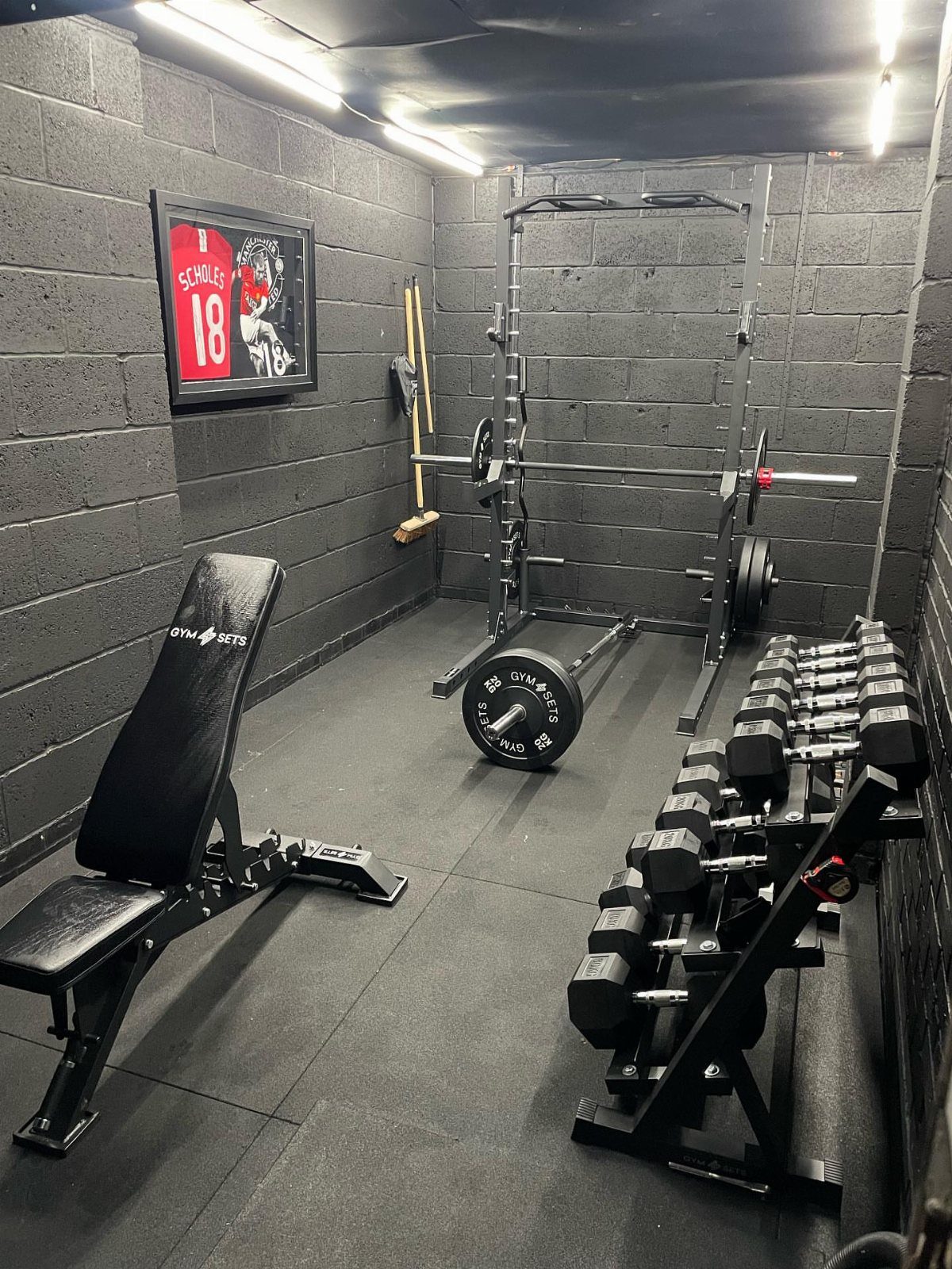 Home gym with weight bench, squat rack, and dumbbells against grey block walls.