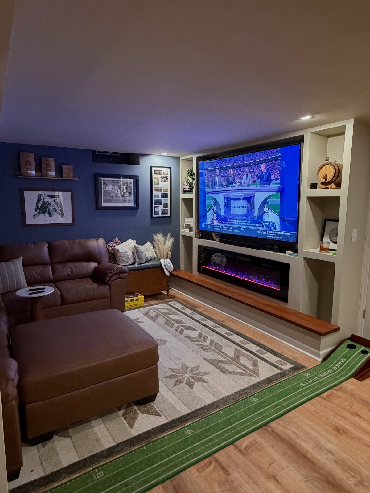 Basement man cave with brown leather sofa, large TV, fireplace, and putting green.