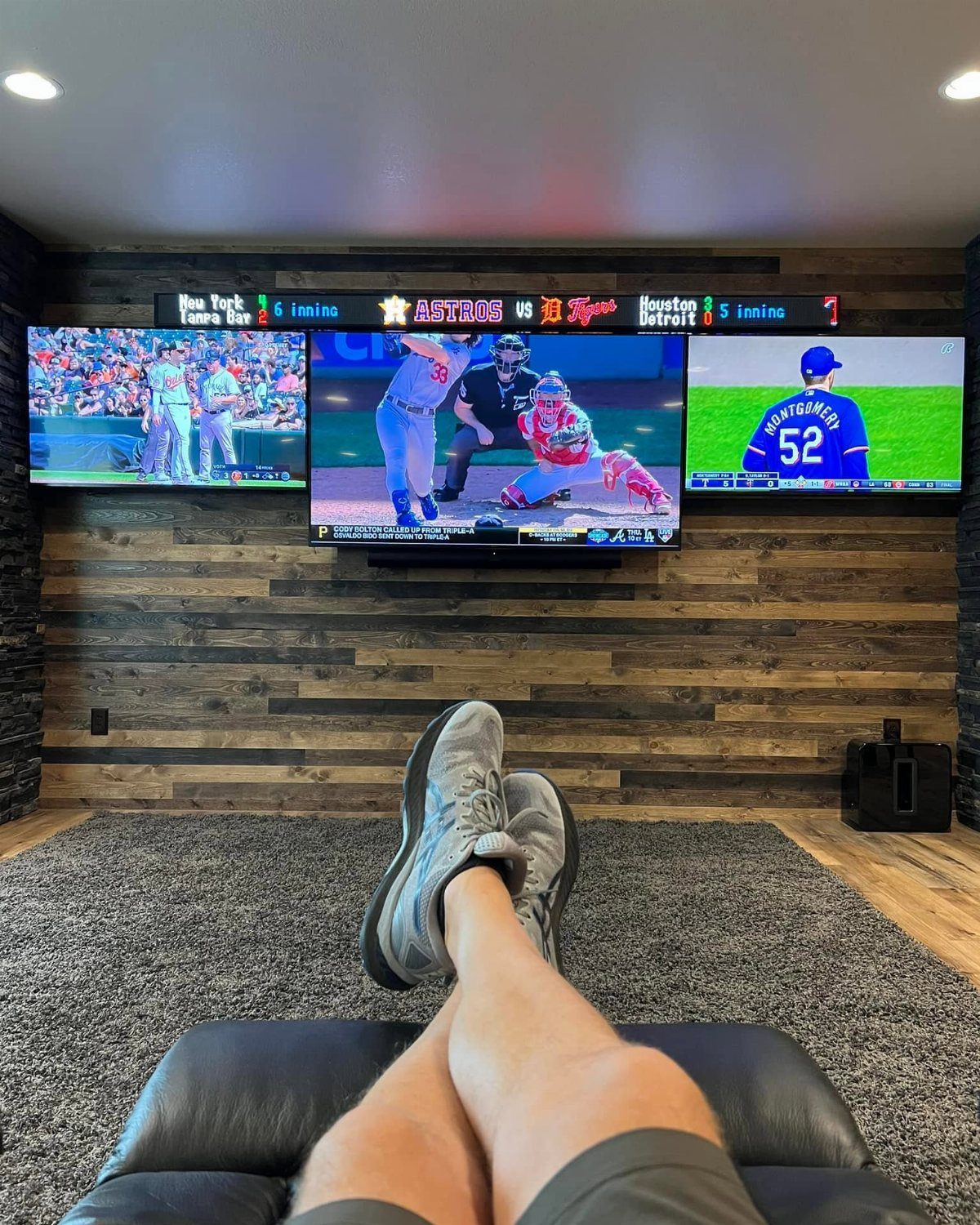 Three televisions showing baseball games on a rustic wood-paneled basement wall.