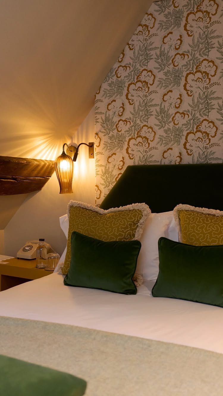 Cozy attic bedroom with green velvet pillows and floral wallpaper under a sloped ceiling.