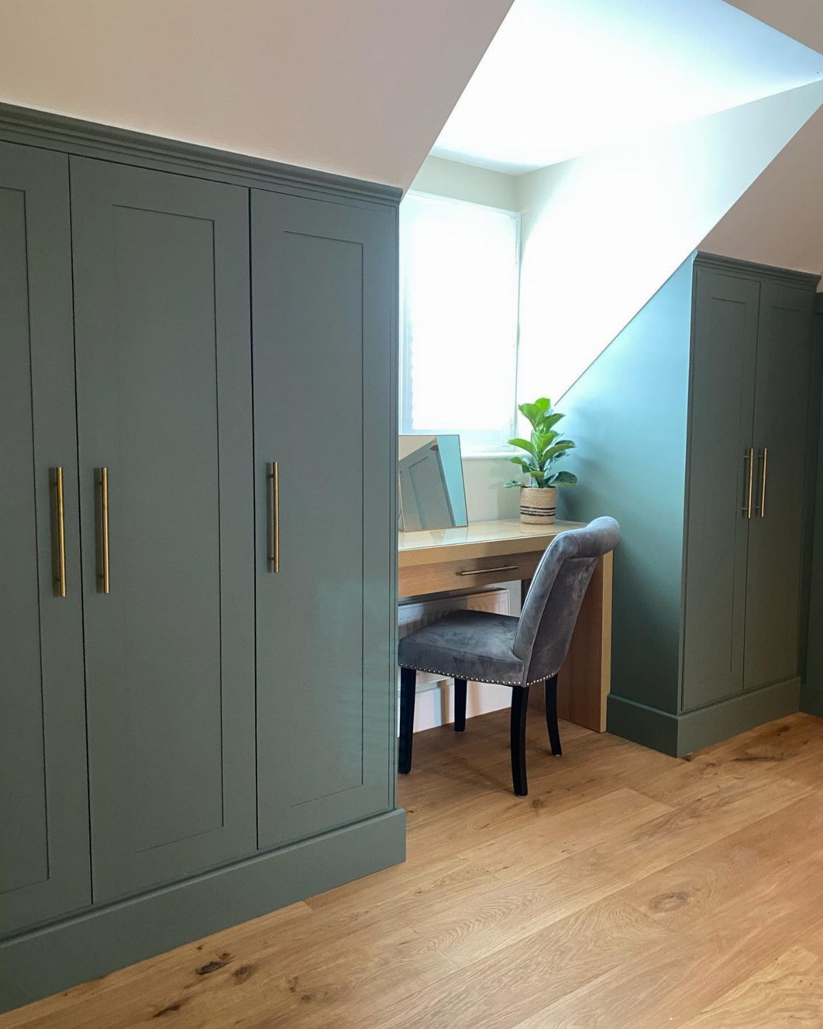 Dormer window illuminating a workspace with teal built-in wardrobes in an attic bedroom.
