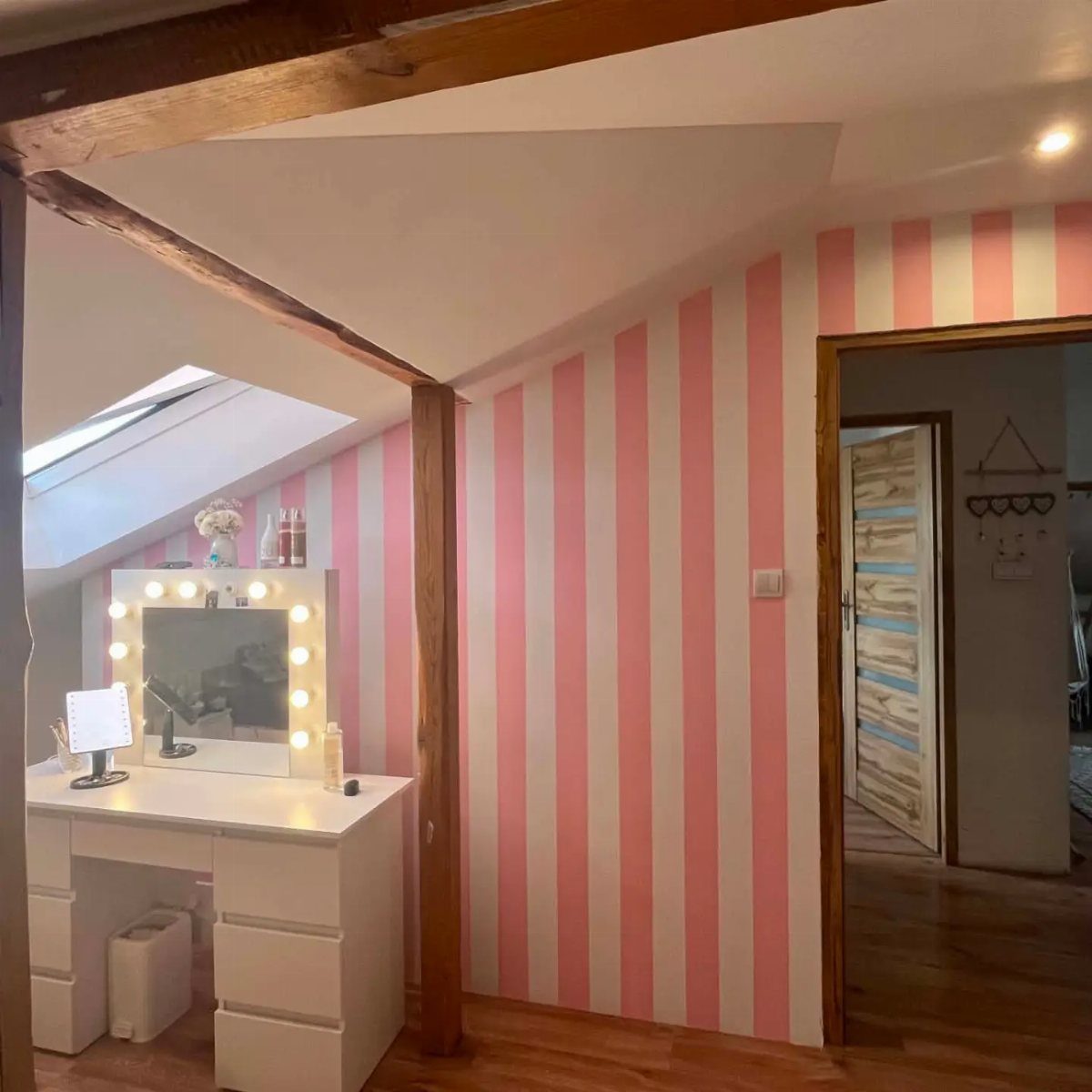 White vanity with lighted mirror against pink striped walls in an attic bedroom.