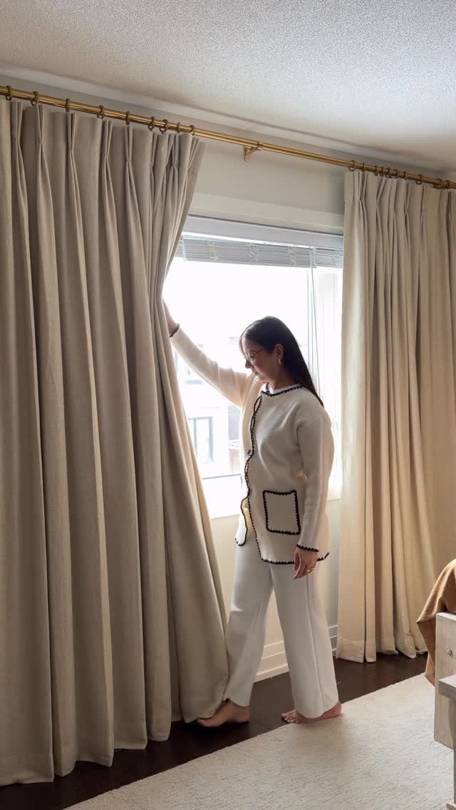 Woman pulling back beige floor-length curtains from a gold rod by a window
