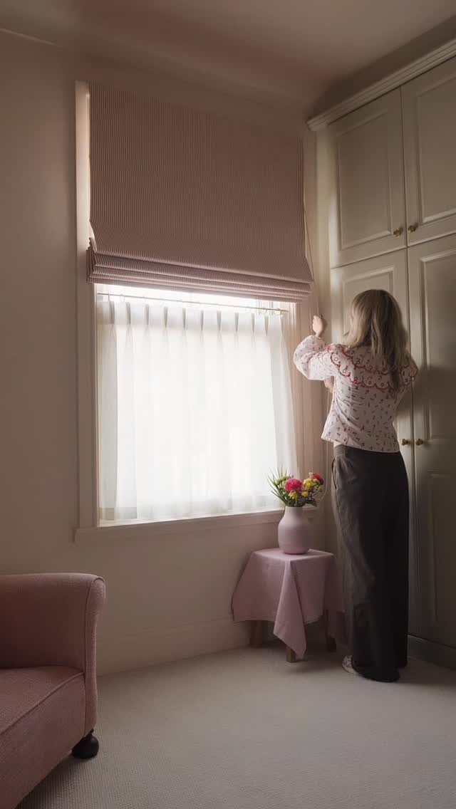 Woman adjusting white sheer cafe curtains under a pink Roman window shade