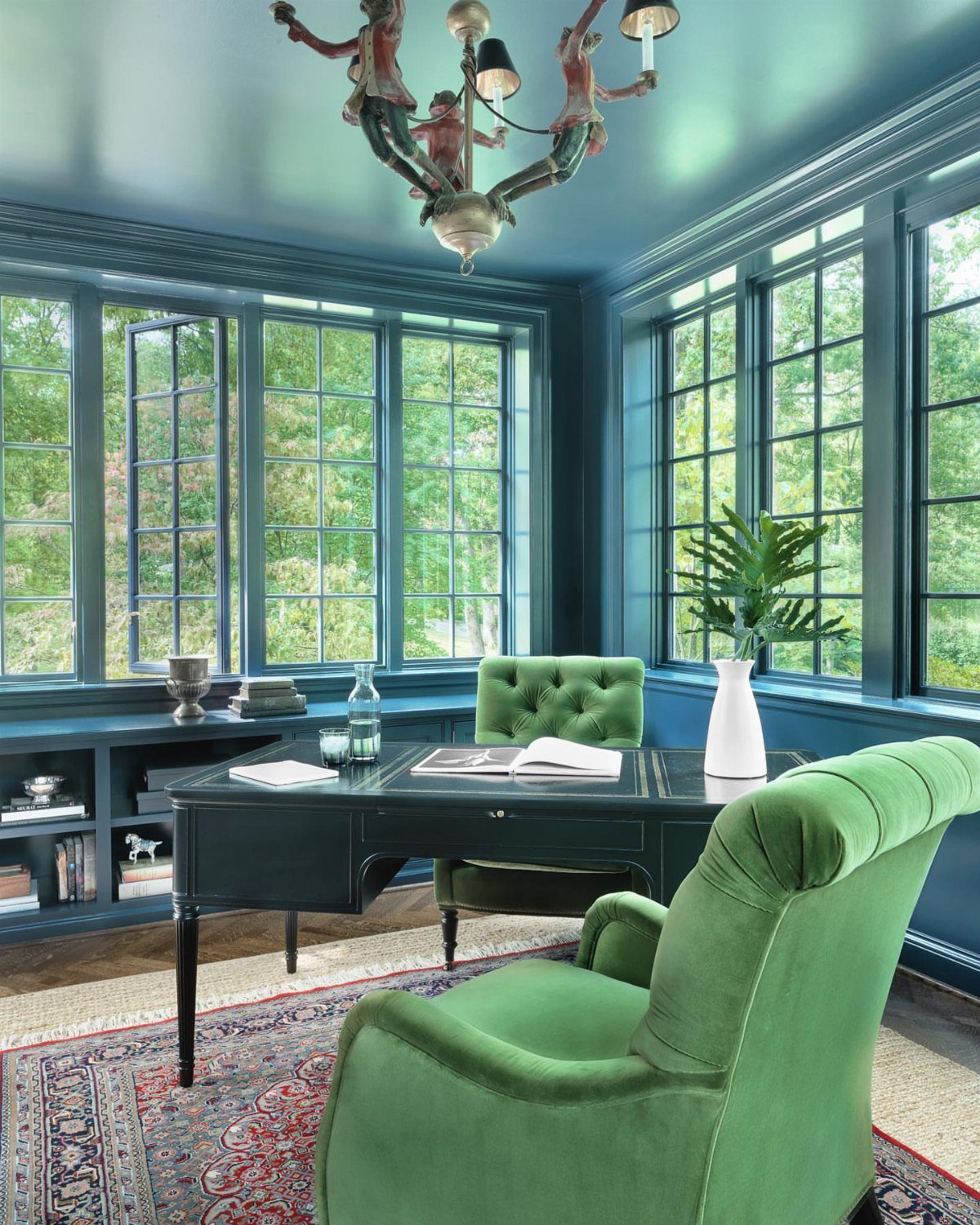 Green velvet tufted armchairs in a dark teal home office with large windows.