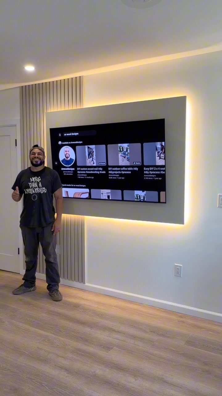 Man standing beside a backlit wall-mounted TV with a wooden slat accent wall.