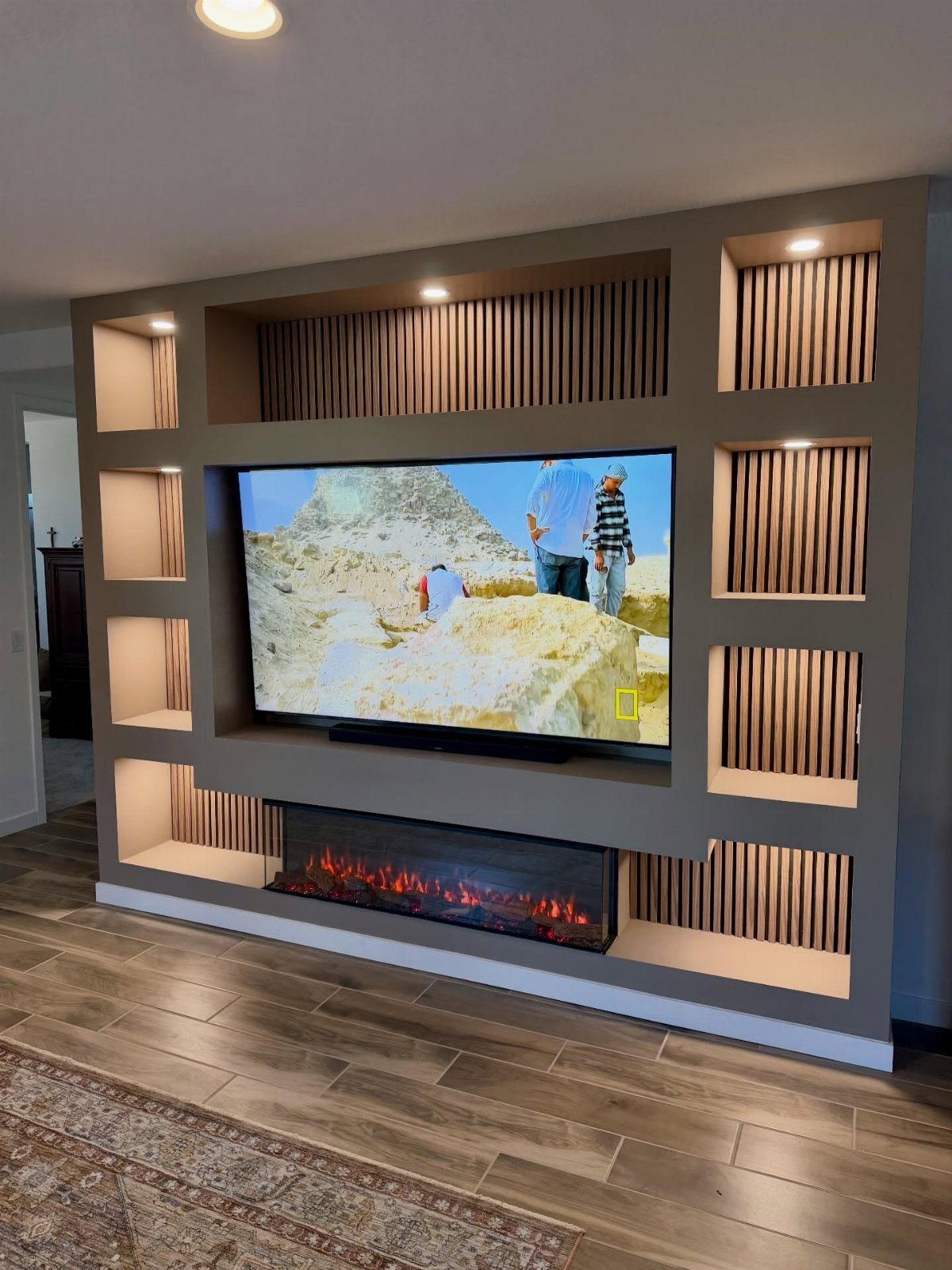 Grey entertainment wall with integrated electric fireplace, mounted TV, and backlit wooden shelving.