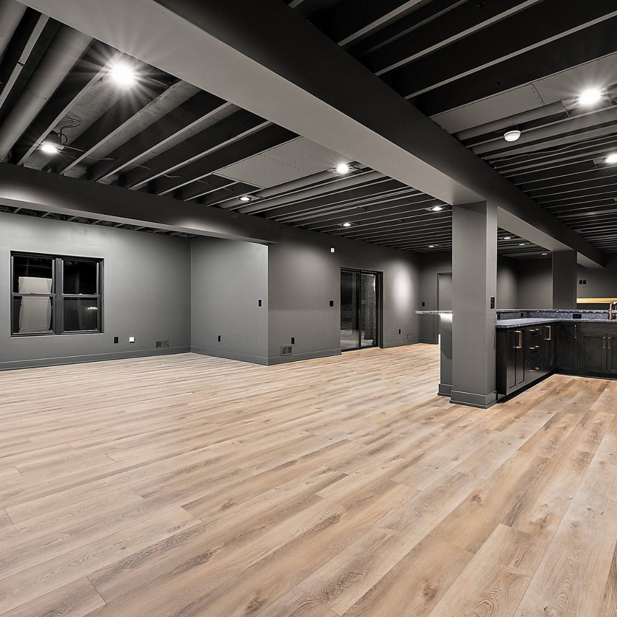 Exposed dark grey basement ceiling with recessed lighting and light wood floors.