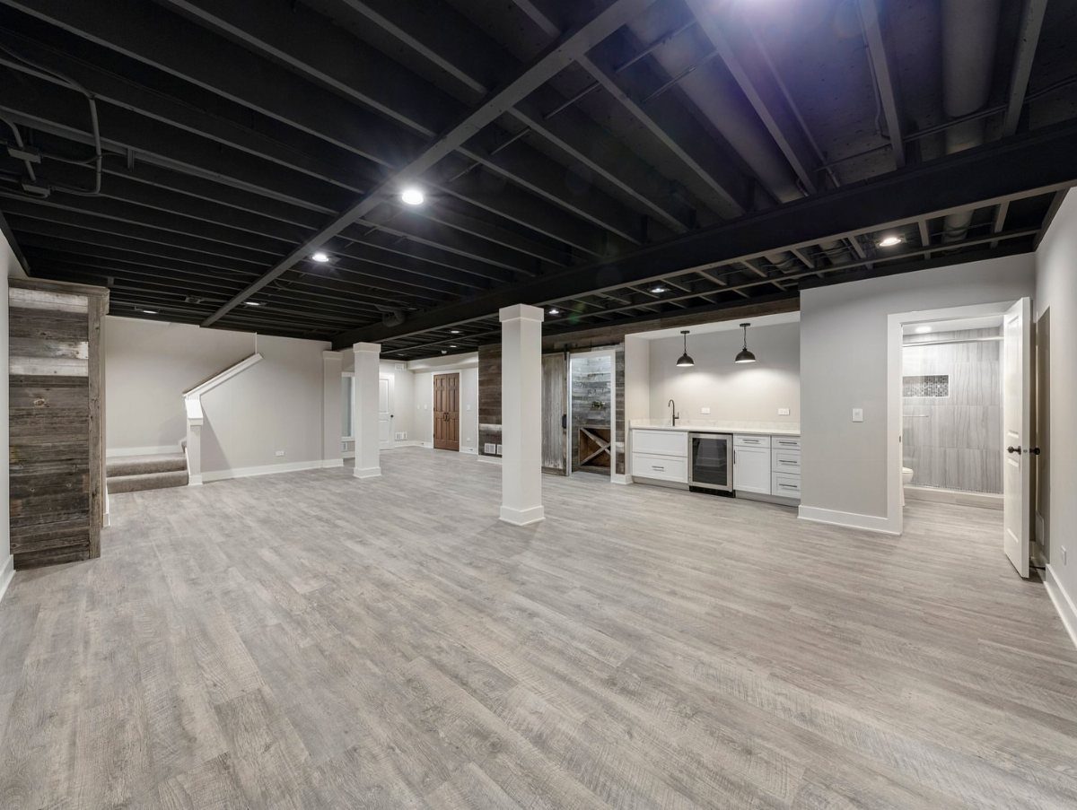 Exposed basement ceiling painted black with recessed lighting and grey wood flooring.