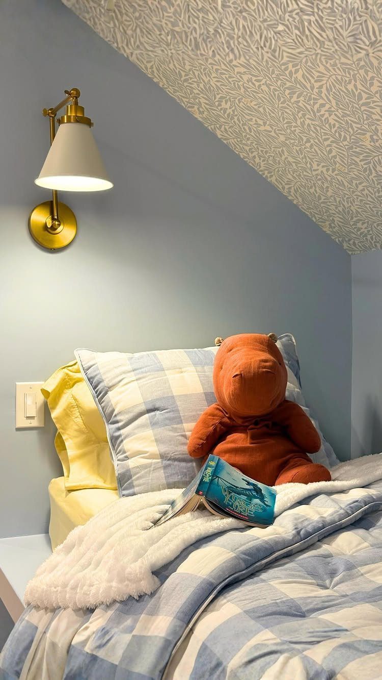 Child's bed with blue checkered linens, stuffed animal, and gold wall lamp in attic.