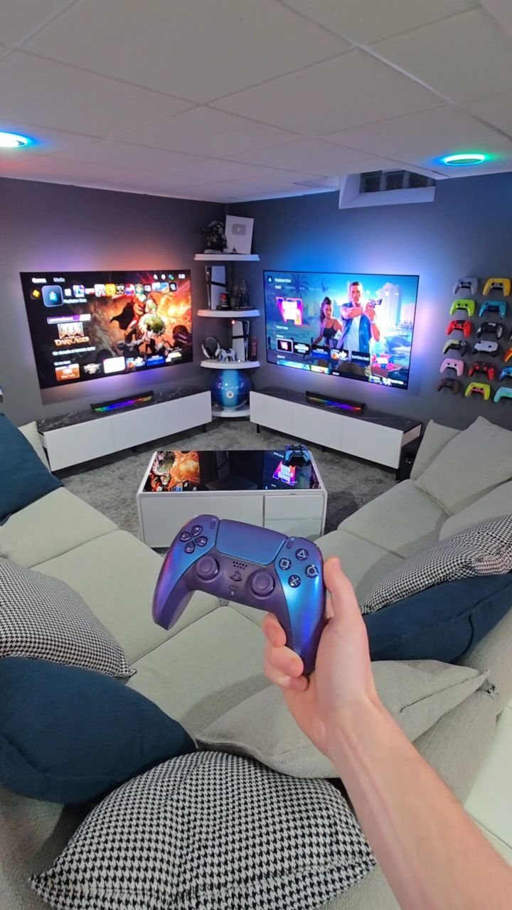 Purple game controller held in front of a basement gaming room with two screens.
