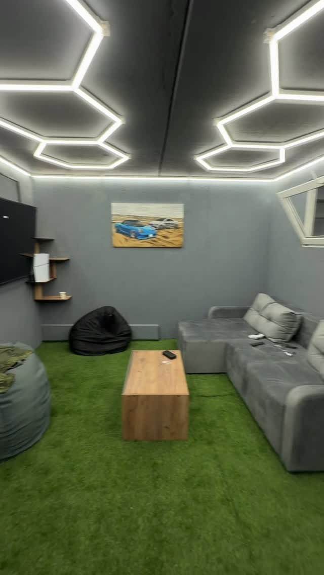 Modern man cave with green grass carpet, grey sofa, and hexagonal ceiling lights.