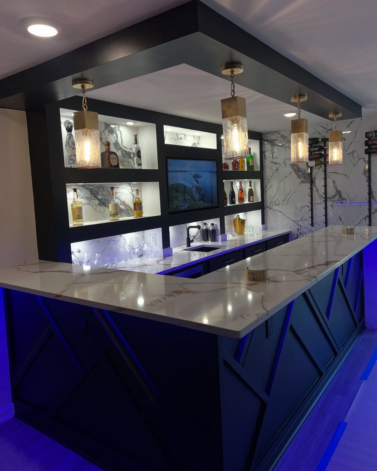 Modern home bar with white marble counters, blue LED lighting, and liquor shelves.