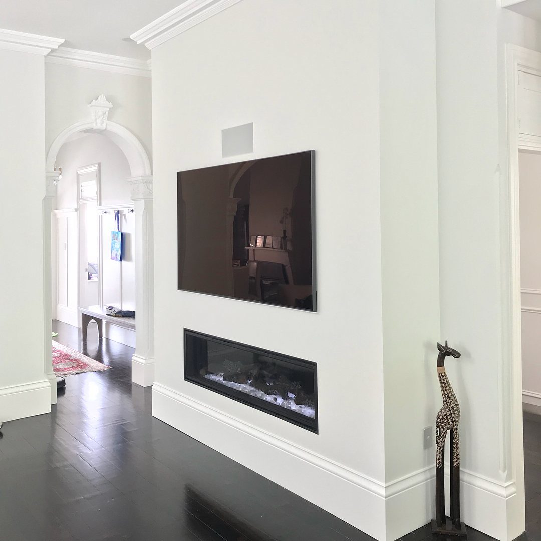 Linear electric fireplace built into a white wall with a television mounted above.