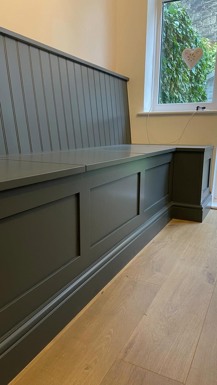 Grey built-in wooden bench with paneled storage base and high backrest by a window.