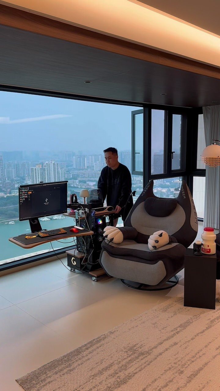 Gaming setup with a PC and ergonomic chair next to large city-view windows.