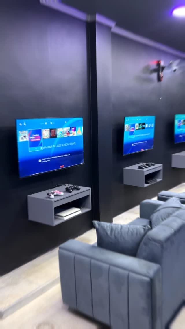 Modern gaming room with wall-mounted TVs, floating shelves with controllers, and grey sofa.