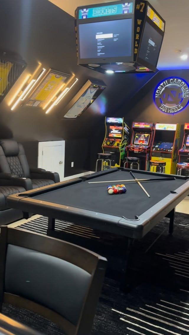 Black pool table in a dark game room with arcade machines and neon lighting.