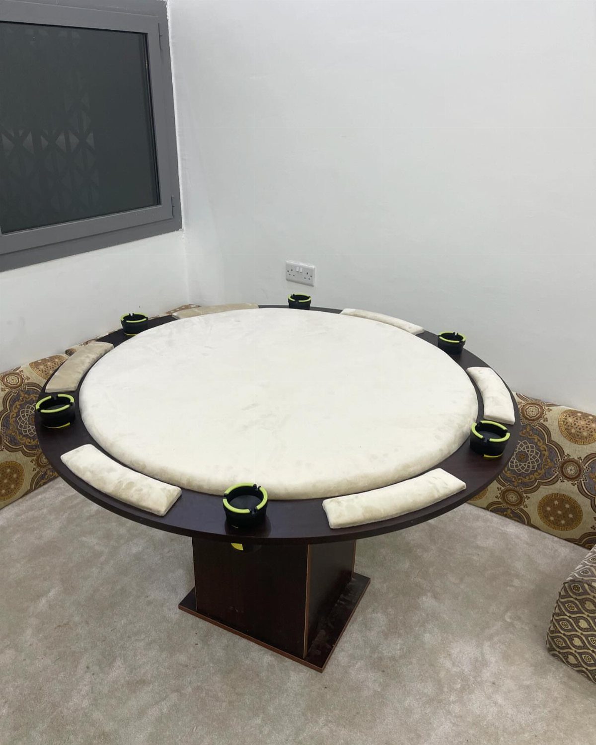 Round gaming table with white padded center and black cup holders in a room.