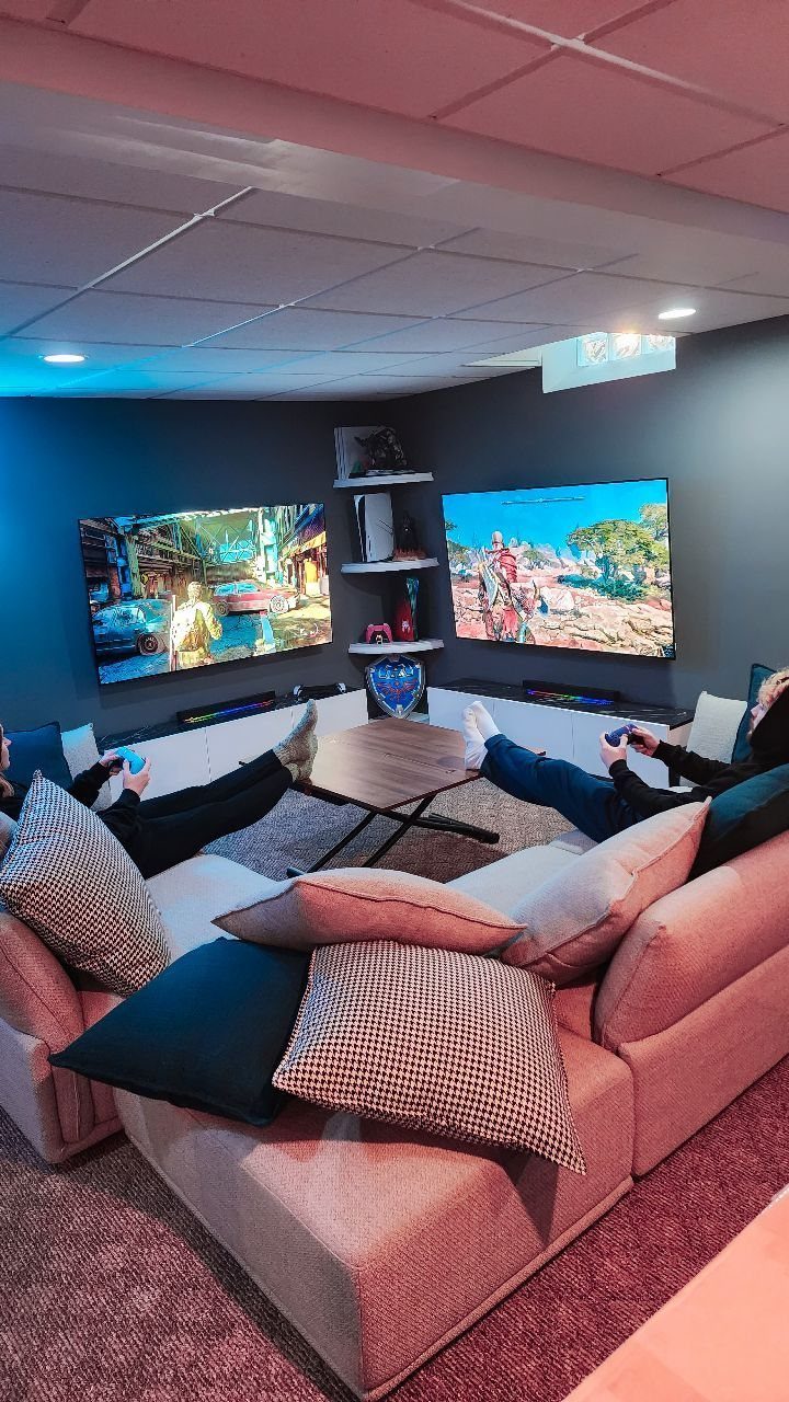 Modern game room with two large screens, grey sectional sofa, and gaming consoles.