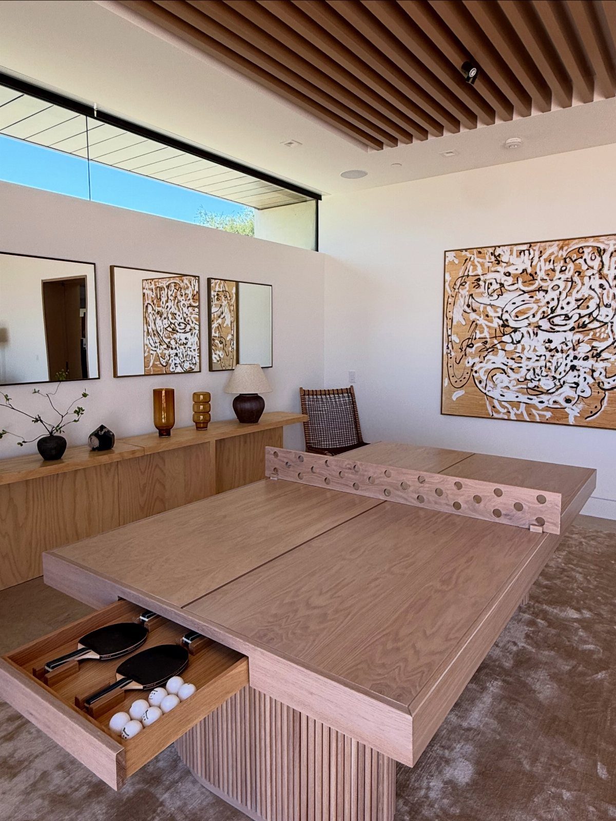 Wooden ping-pong table with paddles and balls in a modern, minimalist room.