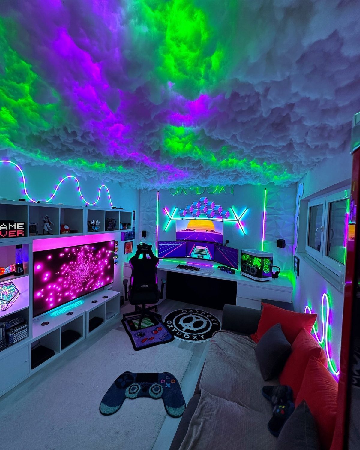 Gaming room with neon lights, cloud ceiling, dual monitors, and a grey sofa.