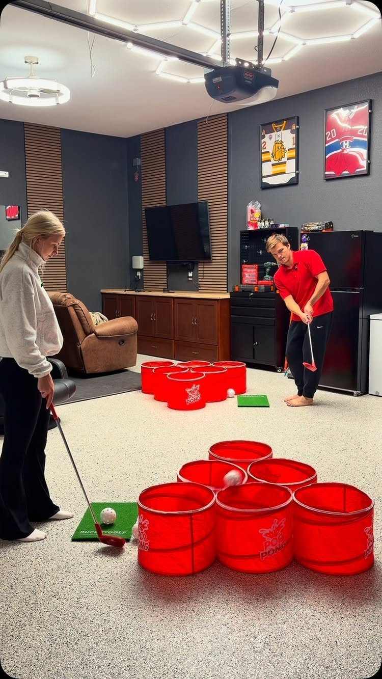 Man and woman playing miniature golf with red buckets in a garage game room.