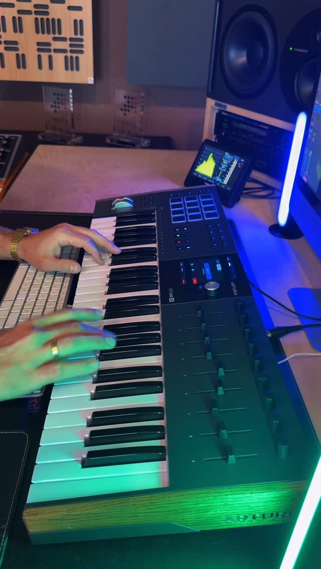 Hands playing a MIDI keyboard controller in a home studio with neon lighting.