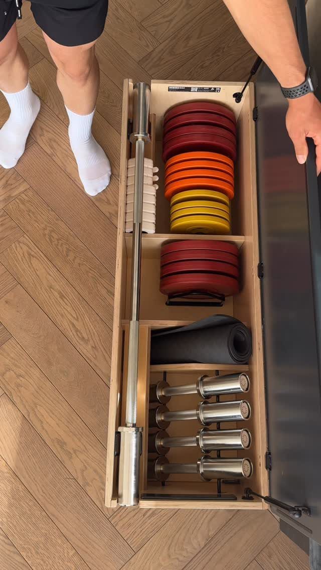 Wooden pull-out drawer organizing a barbell, colorful weight plates, dumbbells, and a mat.
