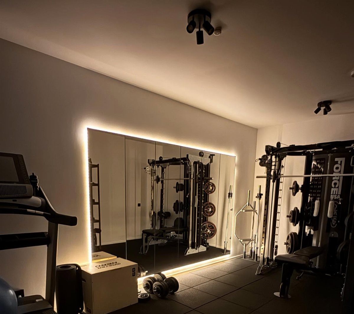 Large backlit wall mirror reflecting gym equipment in a modern home workout room.