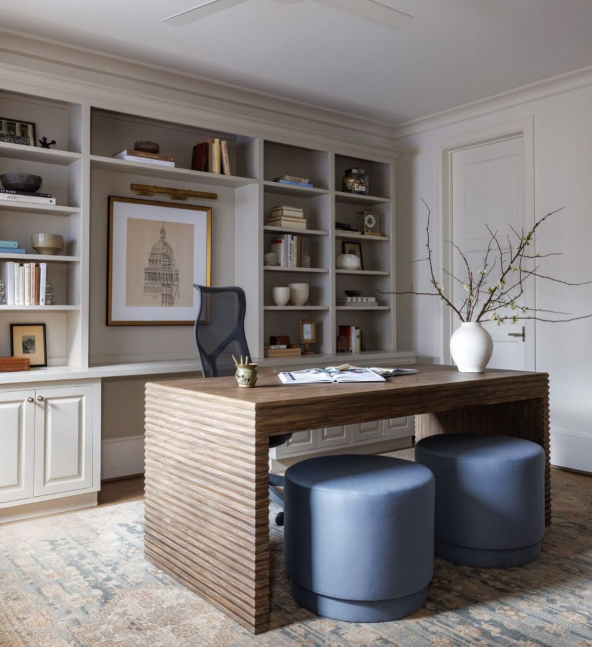 Built-in grey bookshelves and cabinets behind a wooden slatted desk with blue ottomans.