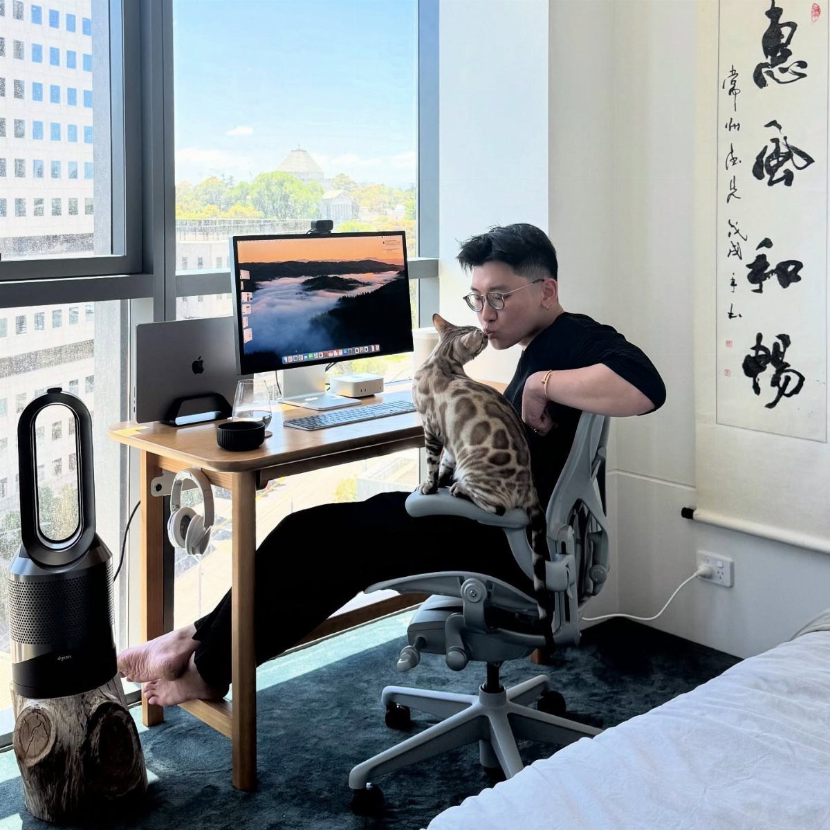 Man sitting in a grey ergonomic office chair at a wooden desk with a cat.