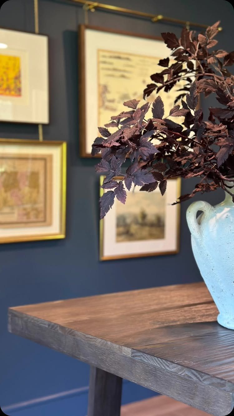 Framed artworks on a dark blue wall behind a wooden table and vase.