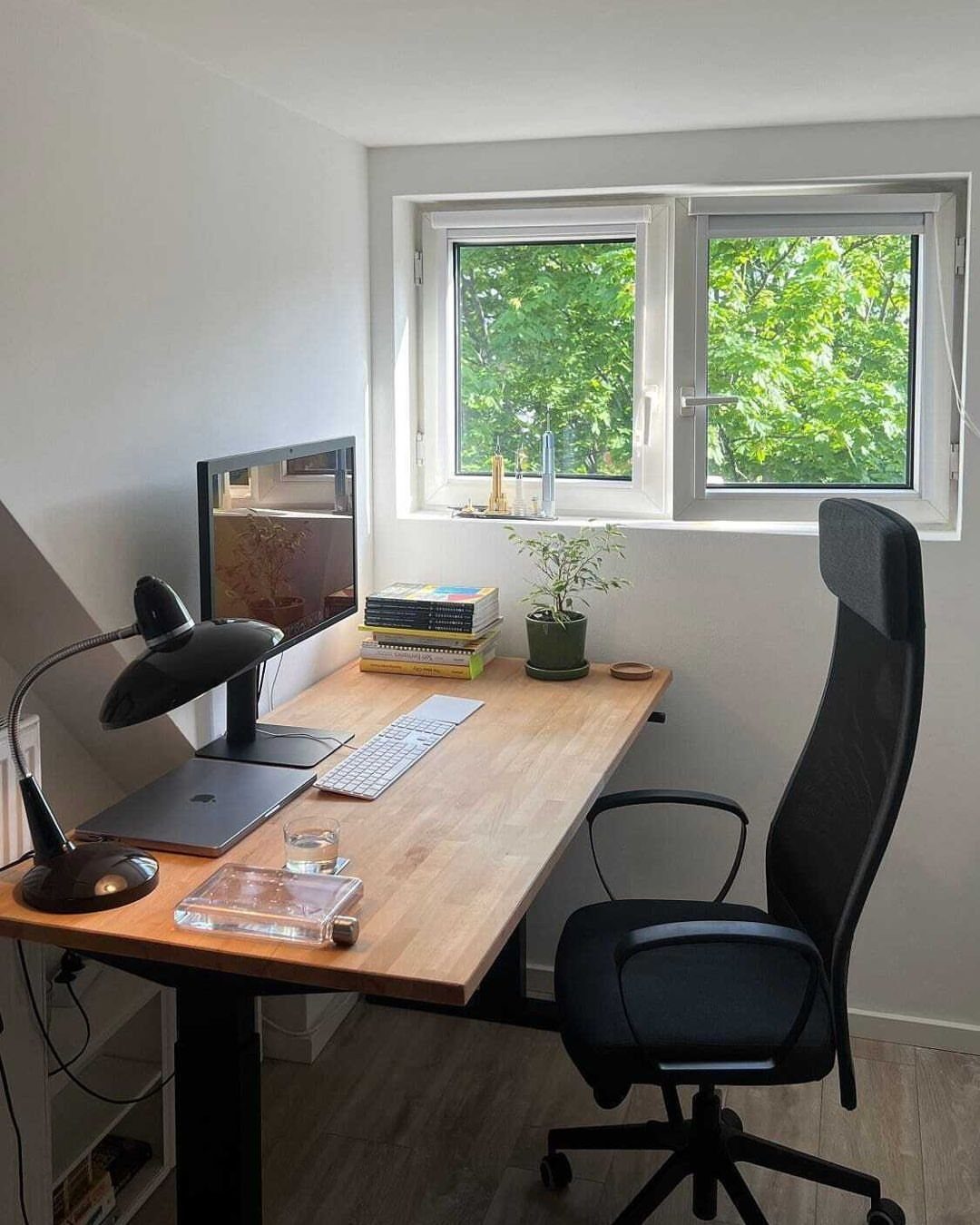 Light wood desk with black ergonomic chair, monitor, and plant by a window