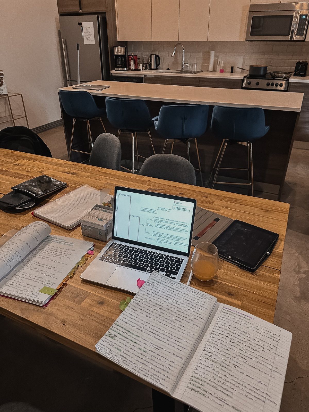 Laptop and open notebooks on a wooden table in an open-concept kitchen area.