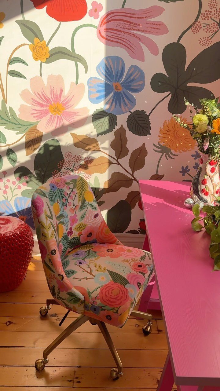 Floral patterned chair and pink desk against a wall with vibrant floral wallpaper.