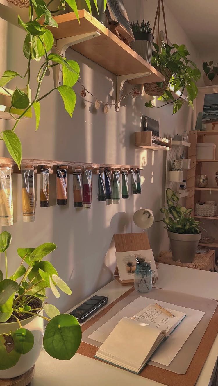 White desk with open notebook, hanging paint tubes, and lush green indoor plants.