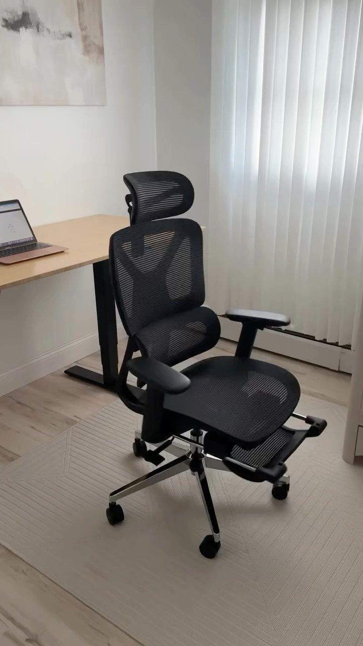 Black ergonomic mesh office chair with footrest next to a wooden desk and laptop